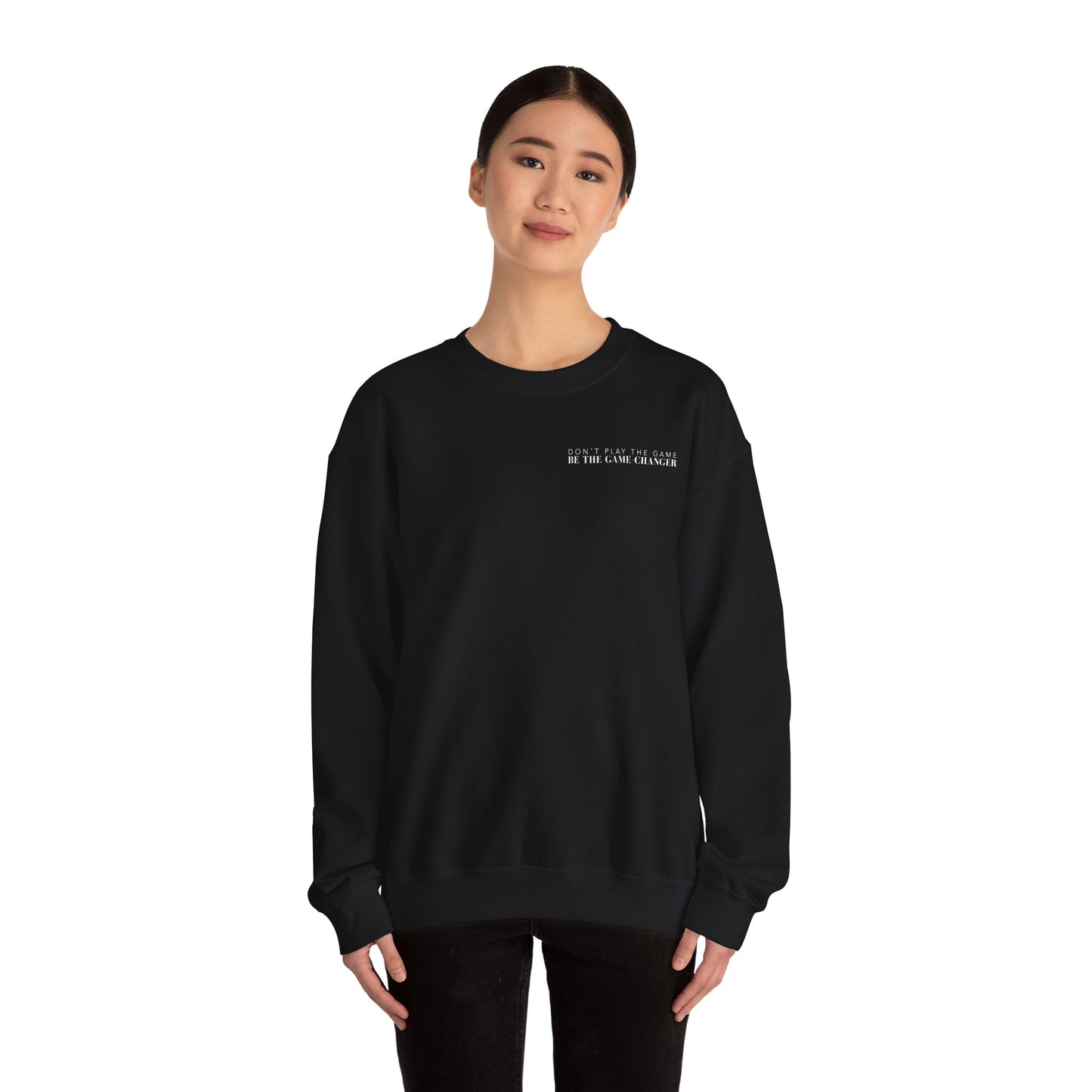 A smiling woman with dark hair and dark eyes wearing black pants and a cozy black crewneck sweater with white letters that say don't play the game be the game-changer over the left chest