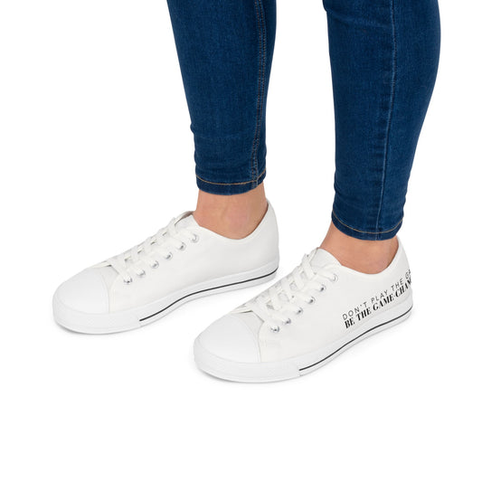 A side view of a woman's legs from the knee down wearing dark wash jeans and women's converse style low top sneaker with don't play the game be the game changer written in black text across the side of the shoe with a black accent line in the leather trim