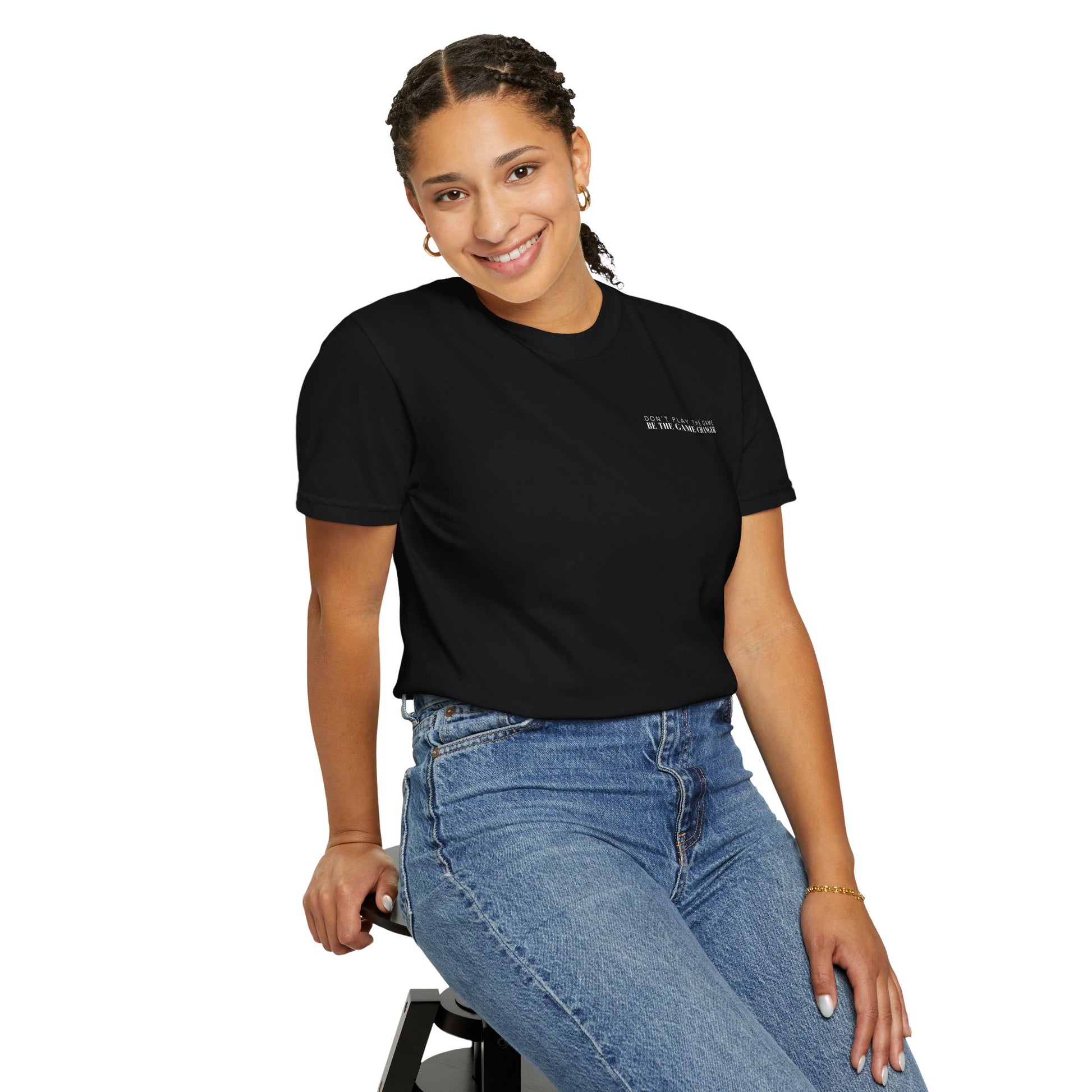 A smiling woman with dark braided hair pulled into a pony tail and dark eyes wearing gold hoops, a gold bracelet, medium wash jeans, and a tucked in short sleeve black t-shirt with white letters that say don't play the game be the game-changer across the left chest