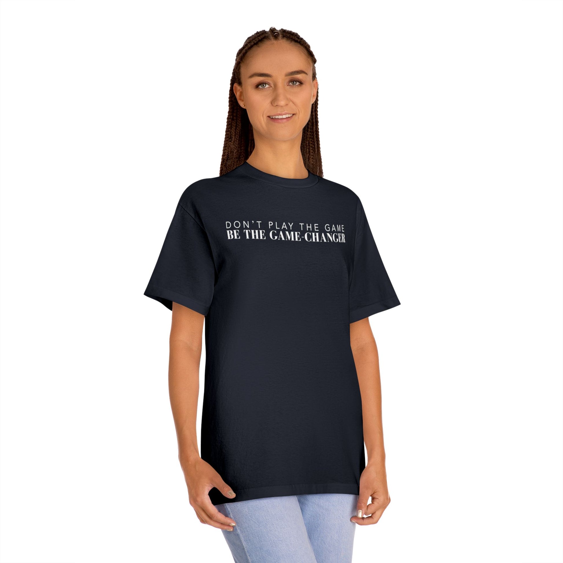A smiling woman with green eyes and brown braids wearing light blue jeans and a short sleeve black t-shirt with white letters that say don't play the game be the game-changer across the chest