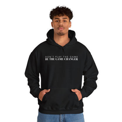 A confident looking man with dark eyes and dark curly hair wearing black hoodie with draw strings, a kangaroo pocket, and white letters that say don't play the game be the game-changer across the chest