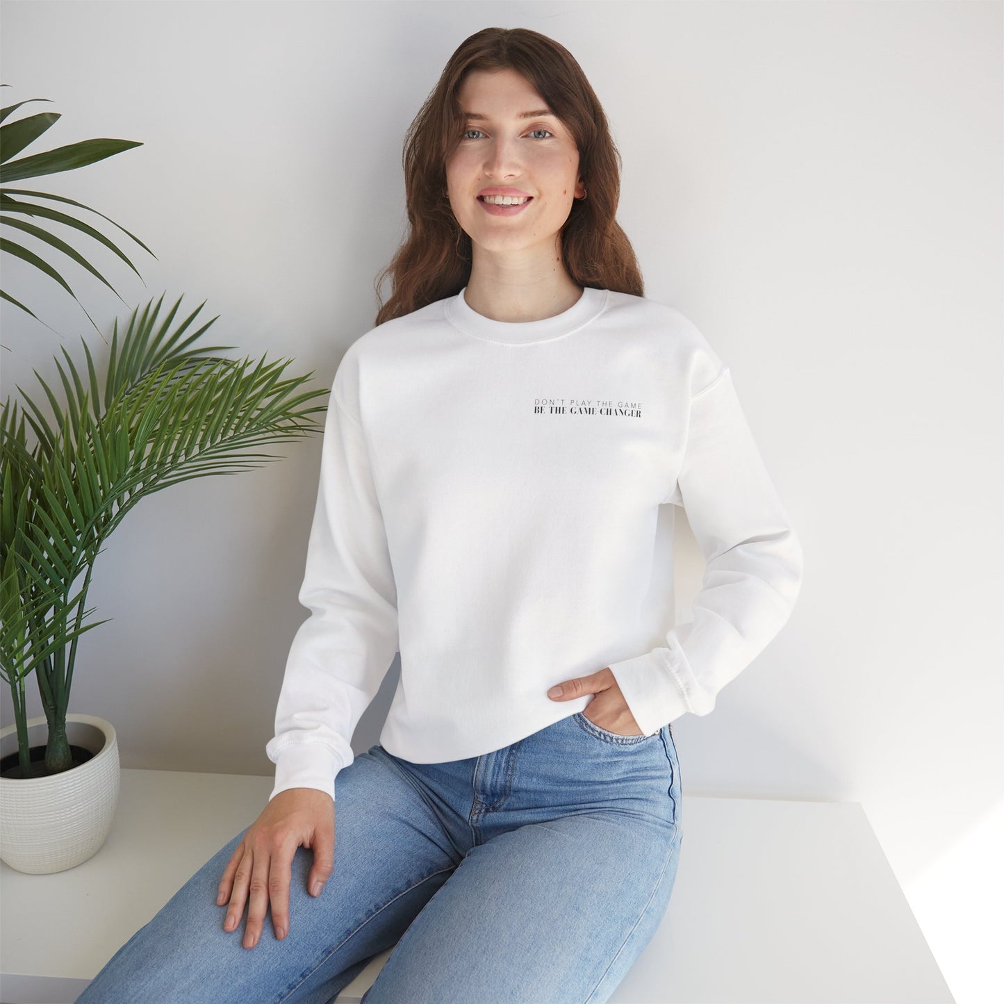 A smiling woman with brown hair and blue eyes seated on a white bench next to a potted palm plant wearing light colored jeans and a cozy white crewneck sweater with black letters that say don't play the game be the game-changer over the left chest 