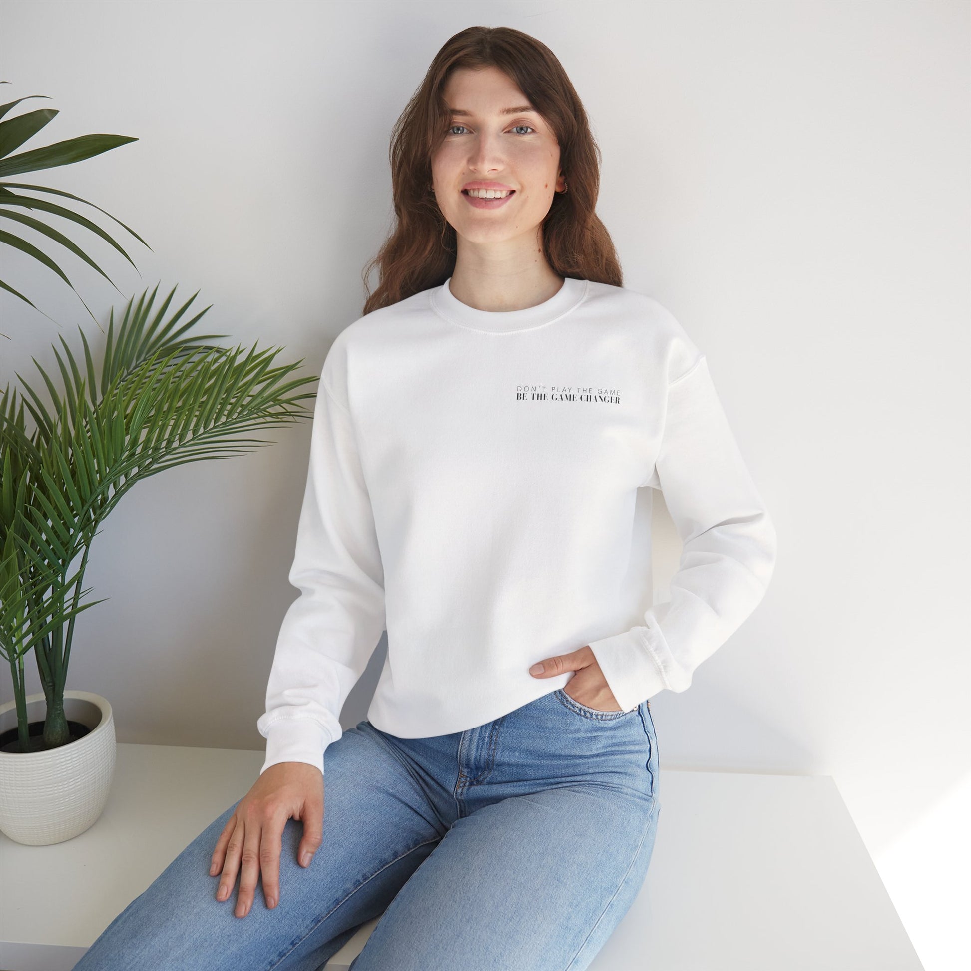 A smiling woman with brown hair and blue eyes seated on a white bench next to a potted palm plant wearing light colored jeans and a cozy white crewneck sweater with black letters that say don't play the game be the game-changer over the left chest 