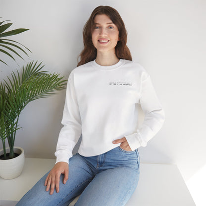A smiling woman with brown hair and blue eyes seated on a white bench next to a potted palm plant wearing light colored jeans and a cozy white crewneck sweater with black letters that say don't play the game be the game-changer over the left chest 