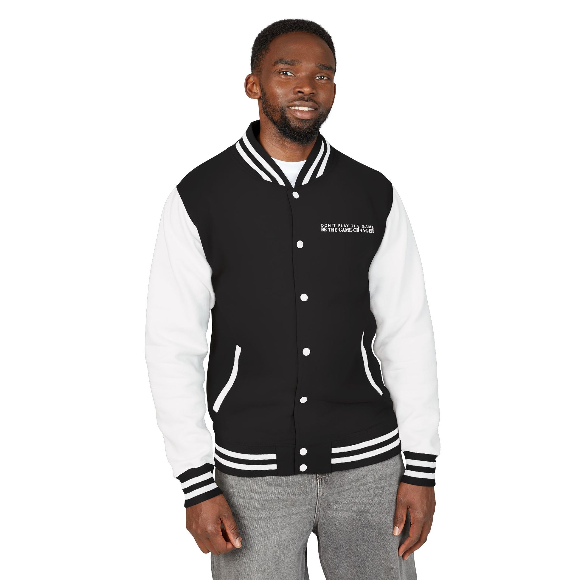 A man with short black curly hair, dark eyes, and facial hair wearing gray jeans and a varsity jacket with white leather sleeves with black and white striped accents, a black fabric body with black and white striped accents on the collar and bottom of the jacket and white pockets and buttons with don't play the game be the game-changer written in white text across the left chest 
