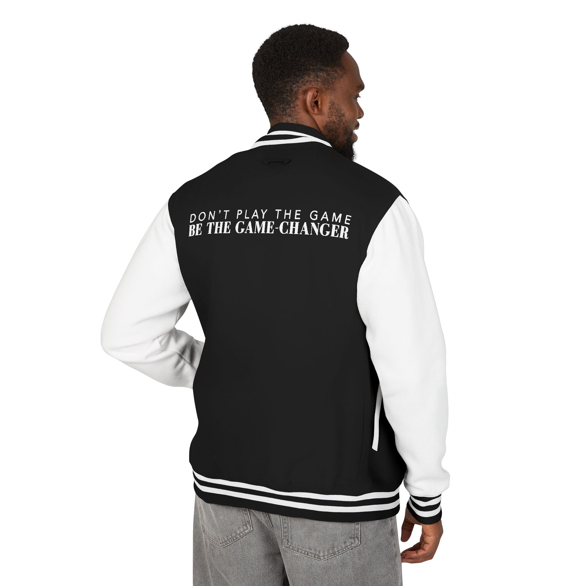 A view from behind a man with short black curly hair wearing gray jeans and a varsity jacket with white leather sleeves with black and white striped accents, a black fabric body with black and white striped accents on the collar and bottom of the jacket with don't play the game be the game-changer written in white text across the back shoulders
