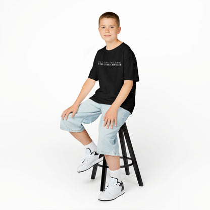 A smiling young boy with brown eyes and short brown hair seated on a black stool wearing jean shorts, white and black sneakers, and a kid's short sleeve black t-shirt with white letters that say don't play the game be the game-changer across the chest