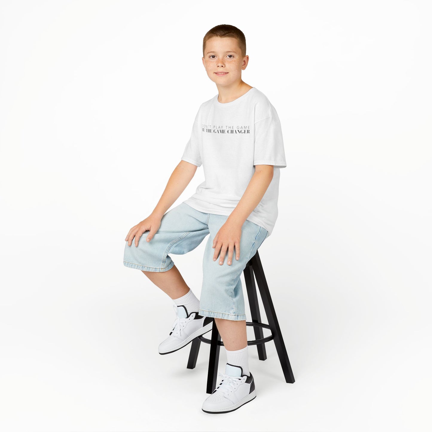 A smiling young boy with brown eyes and short brown hair seated on a black stool wearing jean shorts, white and black sneakers, and a kid's short sleeve white t-shirt with black letters that say don't play the game be the game-changer across the chest