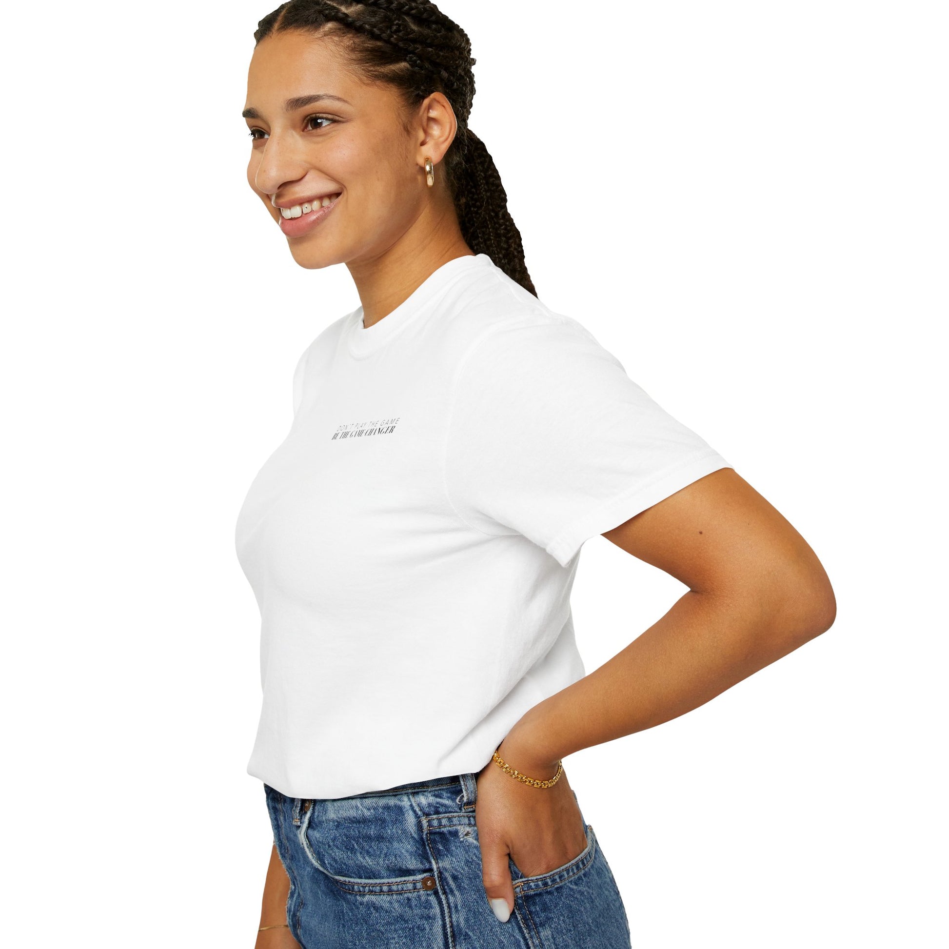 A side view of a smiling woman with dark braided hair pulled back into a pony tail wearing gold hoops, a gold bracelet, and medium wash jeans wearing a tucked in short sleeve white t-shirt with black letters that say don't play the game be the game-changer across the left chest