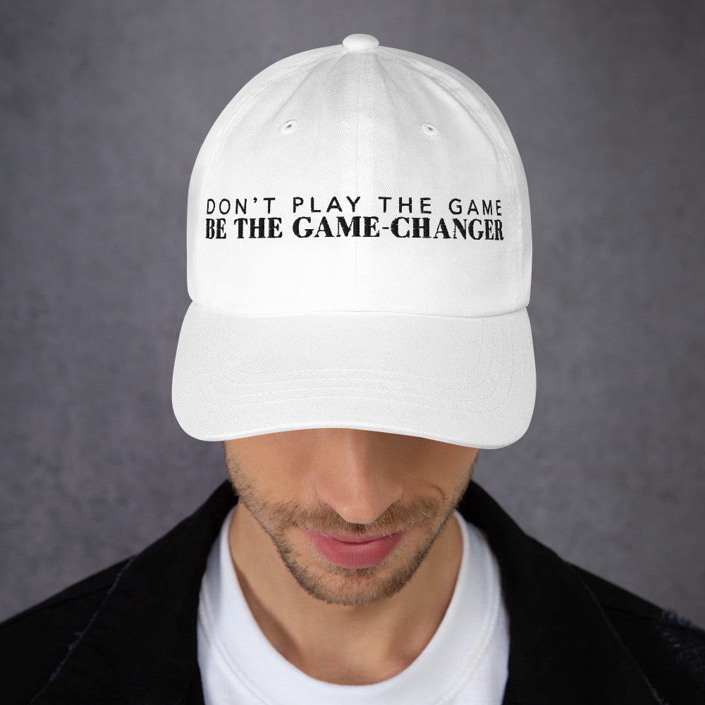 Photo of a man looking down and wearing a white baseball cap that has  black embroidered letters saying don't play the game be the game changer