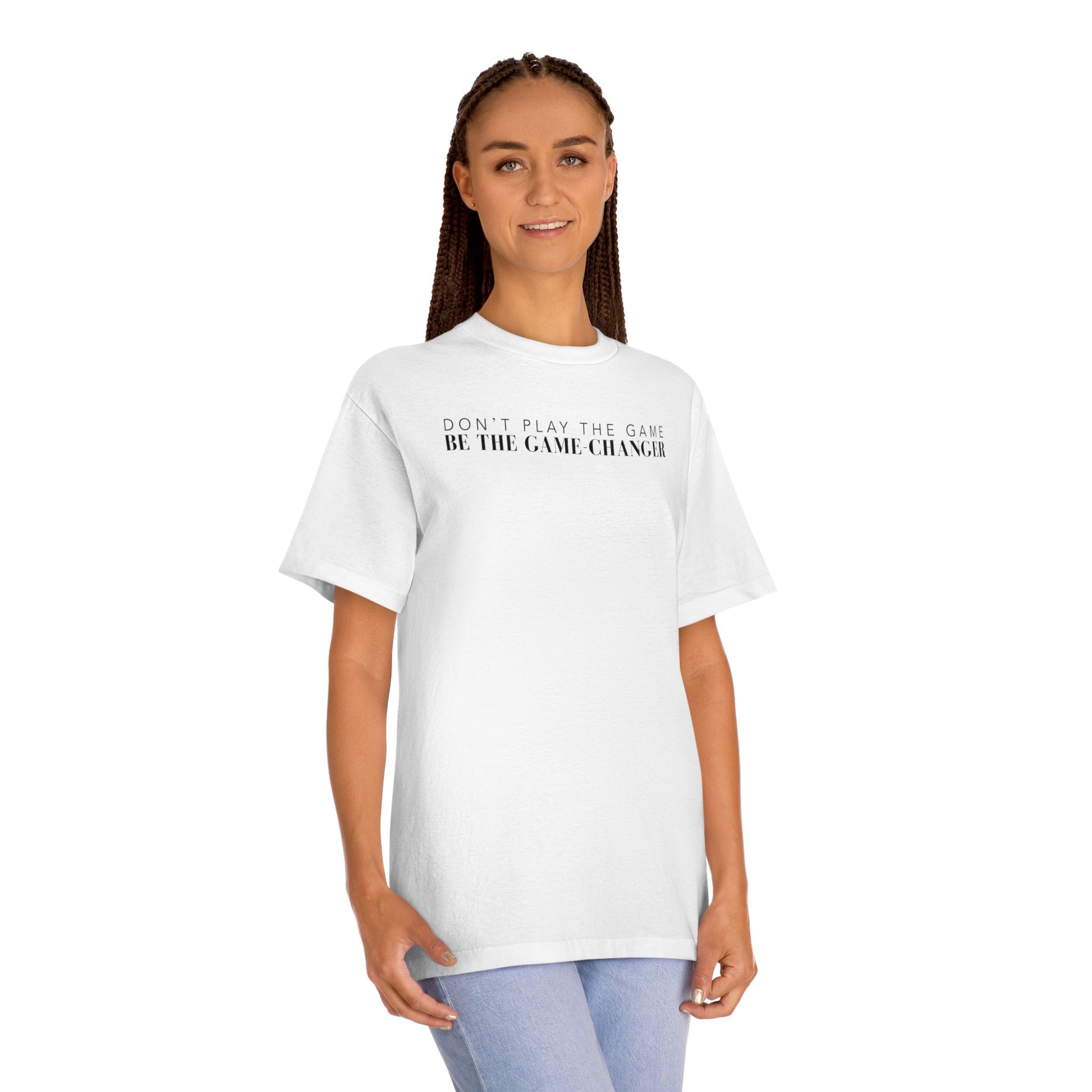 A smiling woman with green eyes and brown braids wearing light blue jeans and a short sleeve white t-shirt with black letters that say don't play the game be the game-changer across the chest