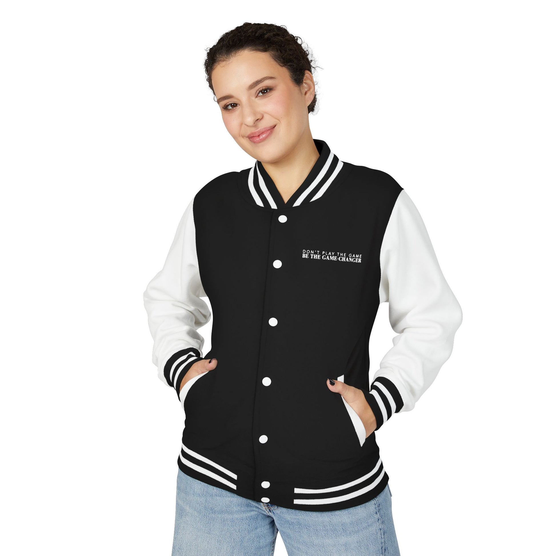 A smiling woman with pulled back dark curly hair, dark eyes, and black nail polish wearing light colored blue jeans and a varsity jacket with white leather sleeves with black and white striped accents, a black fabric body with black and white striped accents on the collar and bottom of the jacket and white pockets and buttons with don't play the game be the game-changer written in white text across the left chest 