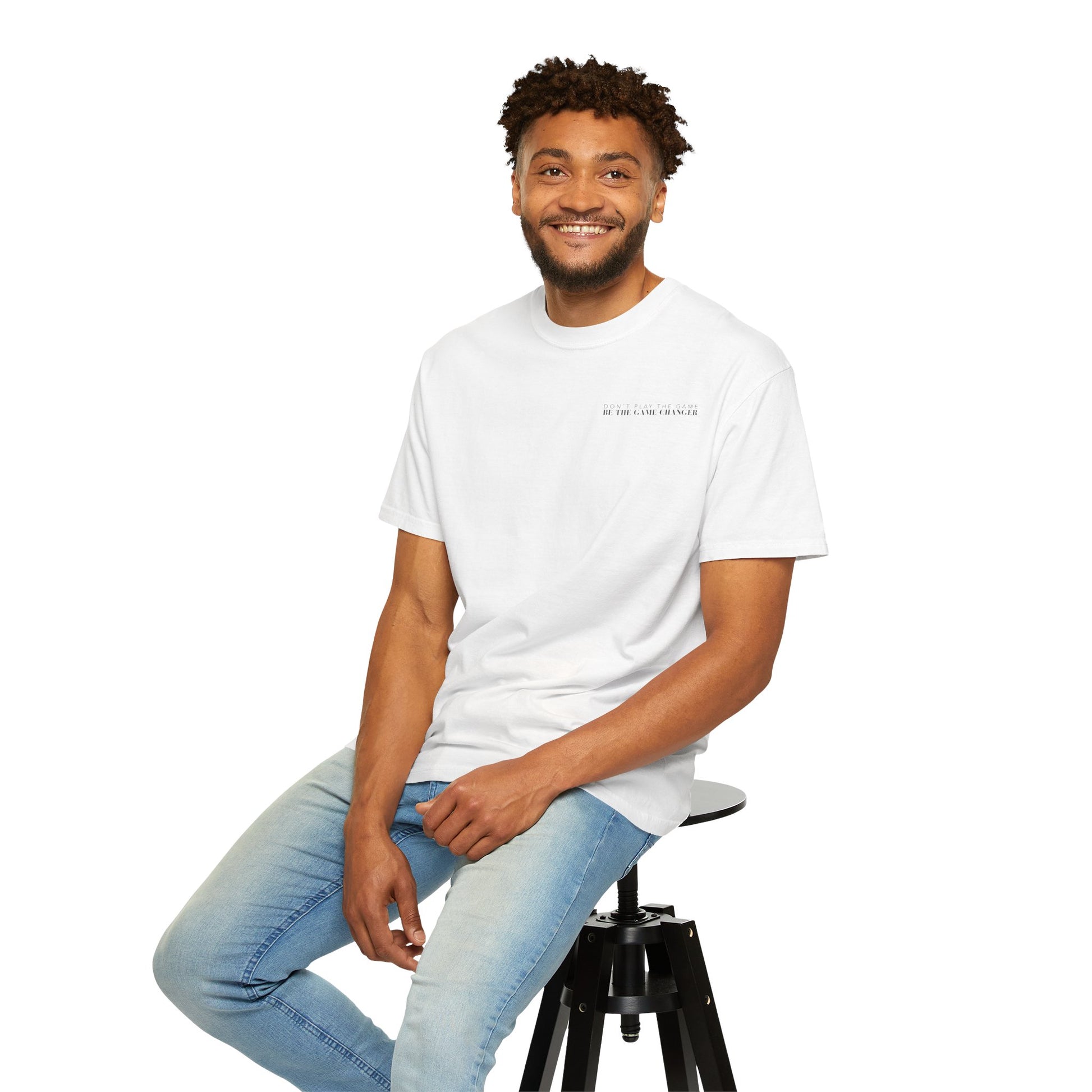 A smiling man with short curly brown hair, dark eyes, and facial hair sitting on a black stool wearing light wash jeans and a short sleeve white t-shirt with black letters that say don't play the game be the game-changer across the left chest