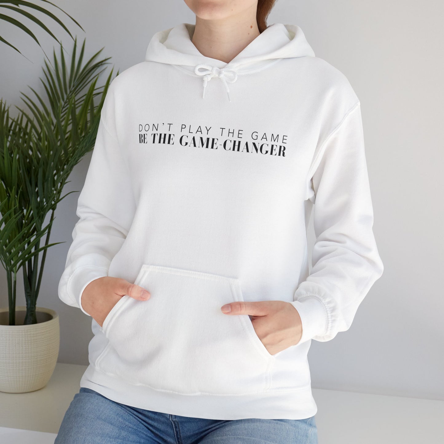 A woman sitting on a white bench next to a potted green palm plant wearing a white hoodie with draw strings, a kangaroo pocket, and black letters that say don't play the game be the game-changer across the chest