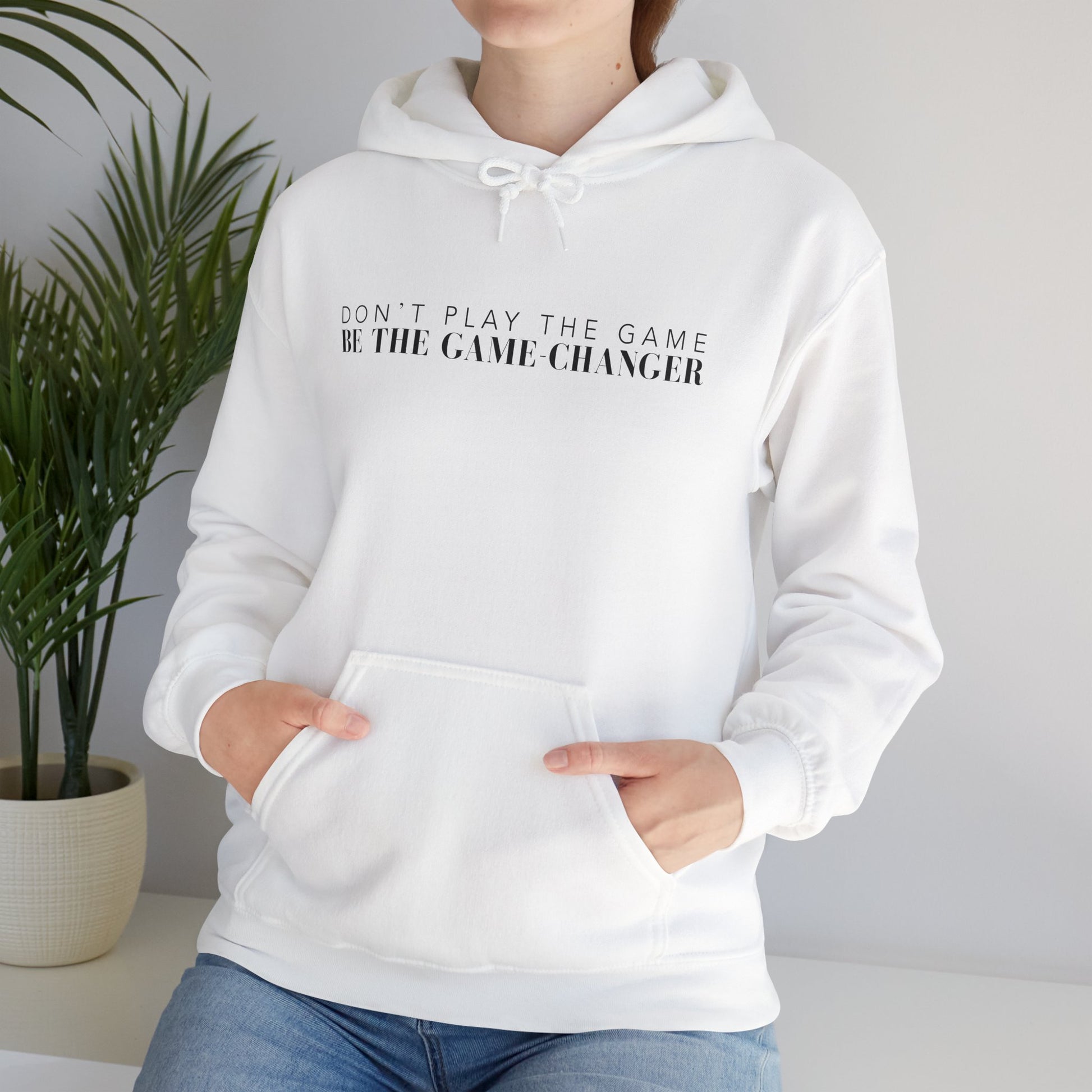 A woman sitting on a white bench next to a potted green palm plant wearing a white hoodie with draw strings, a kangaroo pocket, and black letters that say don't play the game be the game-changer across the chest