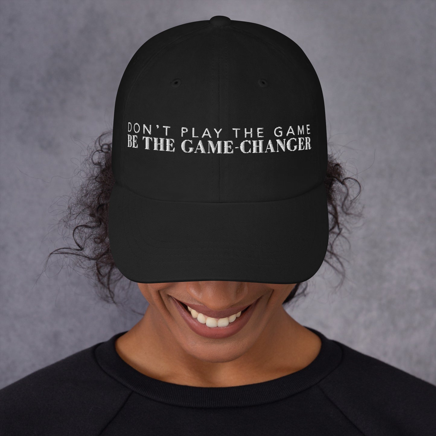 Photo of a smiling woman looking down and wearing a black baseball cap that has white embroidered letters saying don't play the game be the game changer 