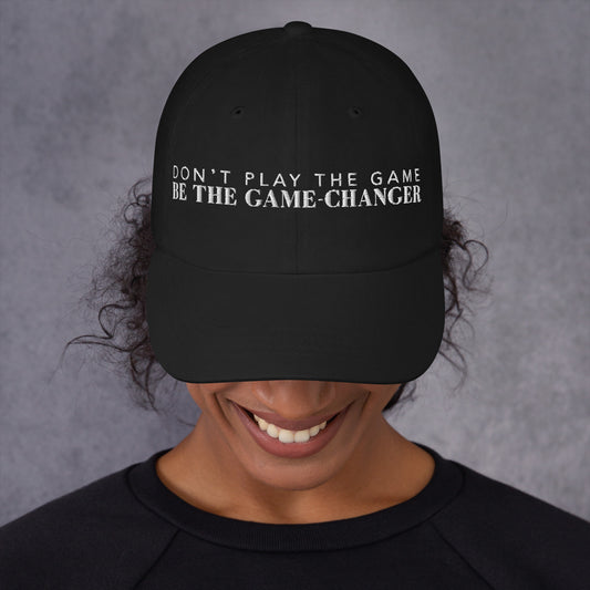 Photo of a smiling woman looking down and wearing a black baseball cap that has white embroidered letters saying don't play the game be the game changer 