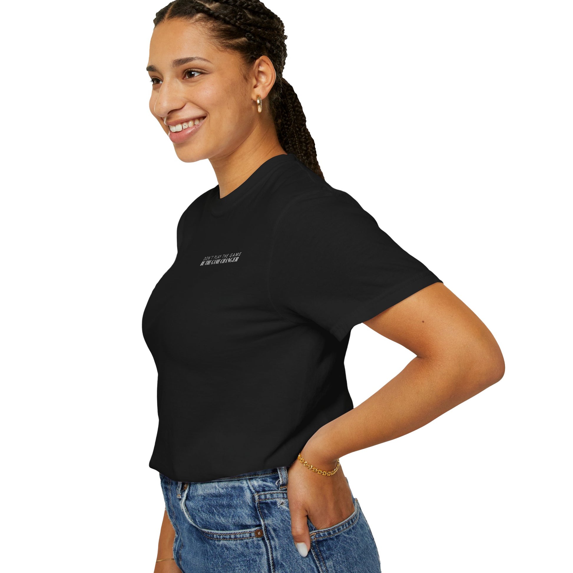 A side view of a smiling woman with dark braided hair pulled back into a pony tail and dark eyes wearing gold hoops, a gold bracelet, medium wash jeans, and a tucked in short sleeve black t-shirt with white letters that say don't play the game be the game-changer across the left chest