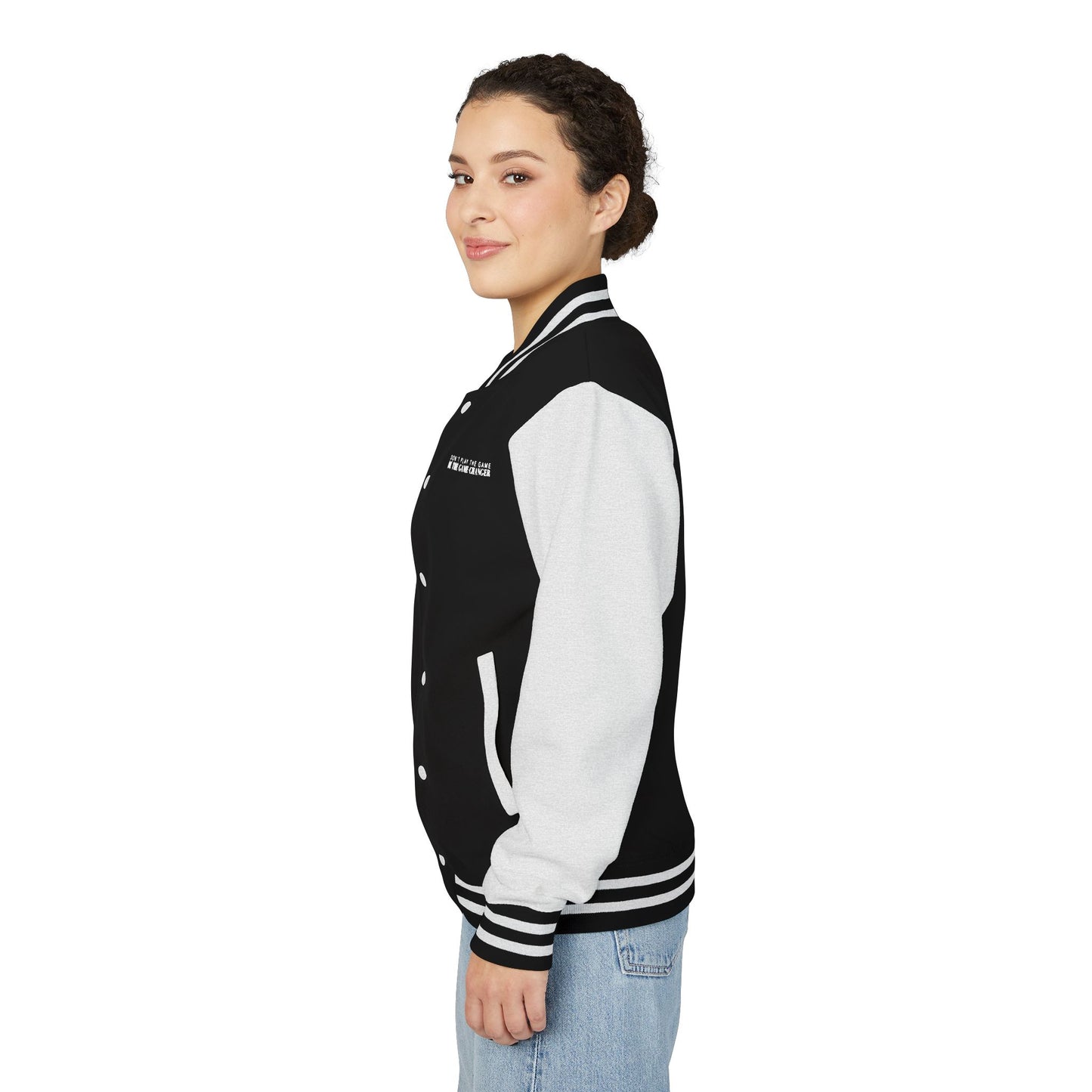The side view of a smiling woman with dark curly hair pulled back into a bun and dark eyes wearing light colored blue jeans and a varsity jacket with white leather sleeves with black and white striped accents, a black fabric body with black and white striped accents on the collar and bottom of the jacket and white pockets and buttons with don't play the game be the game-changer written in white text across the left chest 