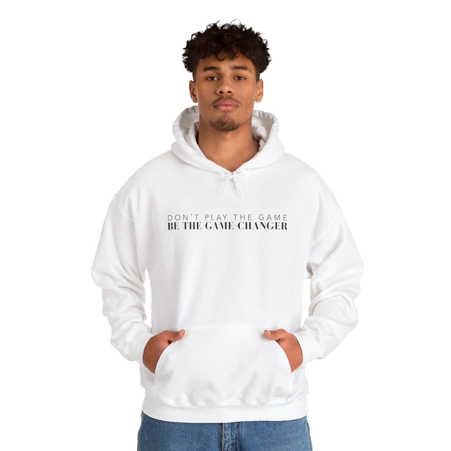 A confident looking man with dark eyes and dark curly hair wearing a white hoodie with draw strings, a kangaroo pocket, and black letters that say don't play the game be the game-changer across the chest