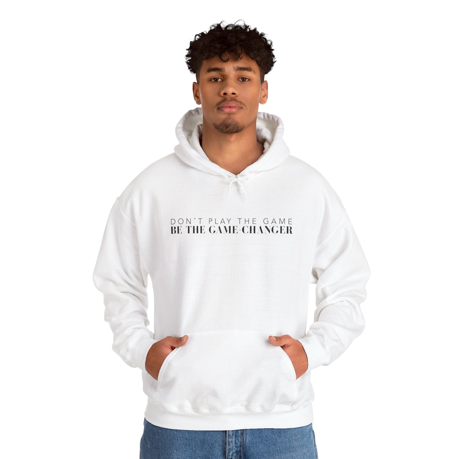A confident looking man with dark eyes and dark curly hair wearing a white hoodie with draw strings, a kangaroo pocket, and black letters that say don't play the game be the game-changer across the chest