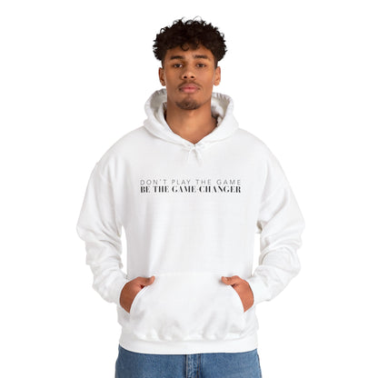 A confident looking man with dark eyes and dark curly hair wearing a white hoodie with draw strings, a kangaroo pocket, and black letters that say don't play the game be the game-changer across the chest