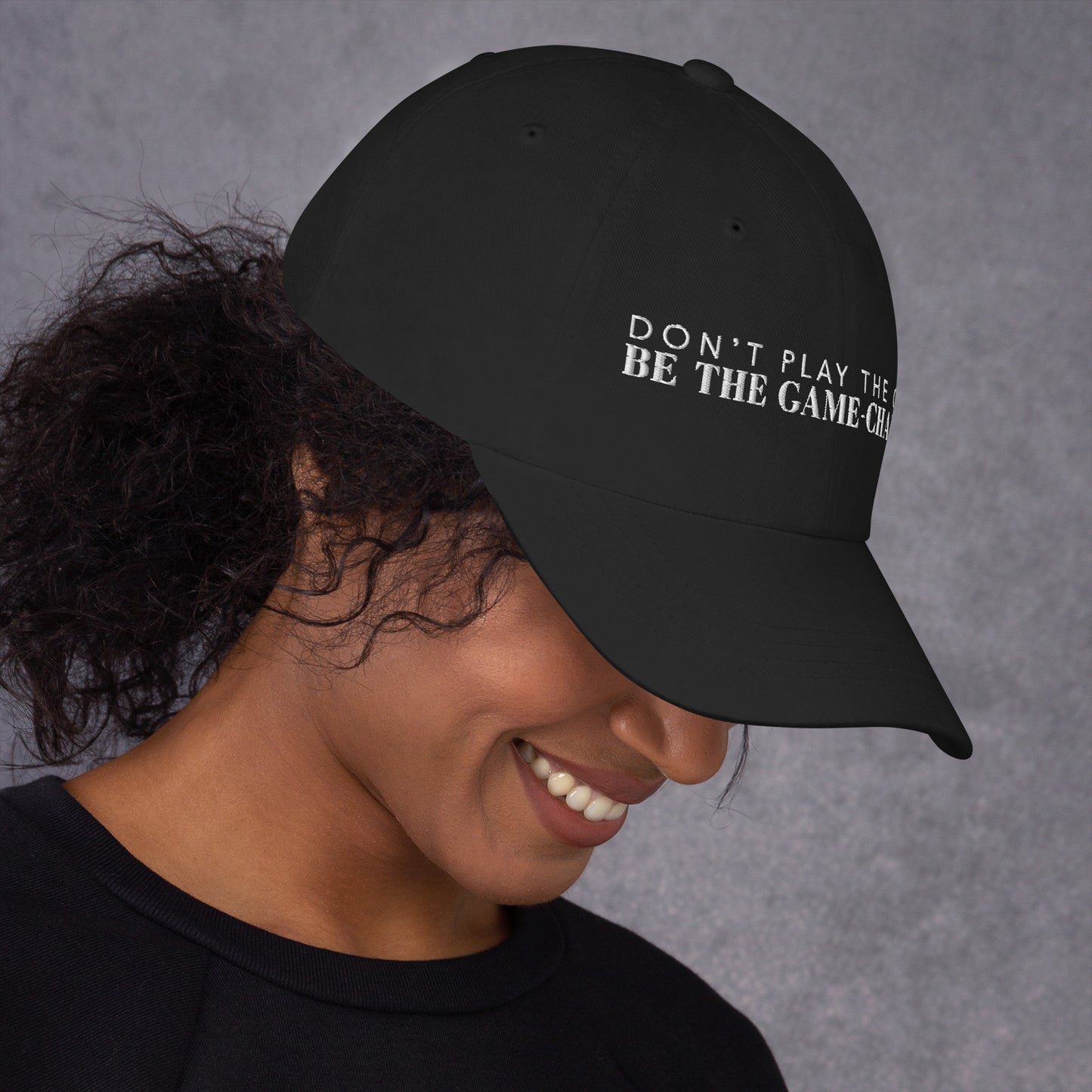 Photo of a woman looking down to the right and wearing a black baseball cap that has white embroidered letters saying don't play the game be the game changer 