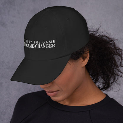 Photo of a woman looking down to the left and wearing a black baseball cap that has white embroidered letters saying don't play the game be the game changer 
