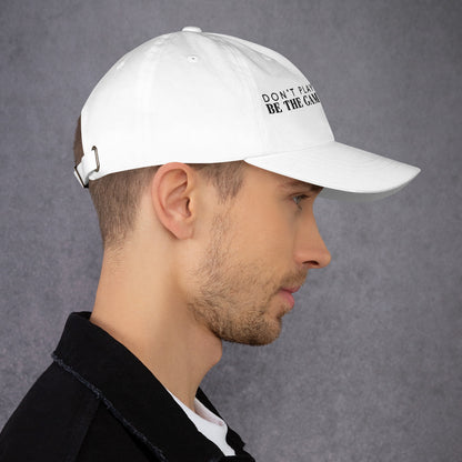 Photo of a man looking to the right and wearing a white baseball cap that has  black embroidered letters saying don't play the game be the game changer