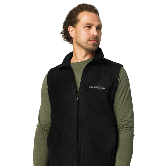 A confident man with brown curly hair pulled back into a bun, light eyes, and facial hair looking to the right and wearing a green long sleeve shirt and A Black fleece men's vest with don't play the game be the game changer written in small text across the left chest