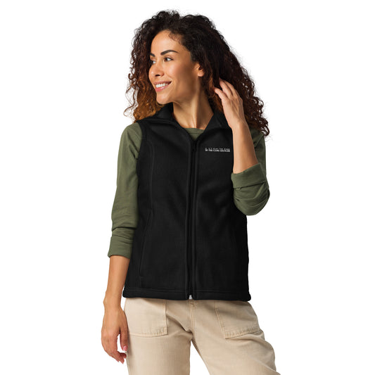 A smiling woman with curly hair with a brown ombre and dark eyes wearing khaki pants, a long sleeve green shirt, and a black Columbia fleece vest with don't play the game be the game-changer written in small white text across the left chest 